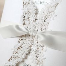 White satin lace tied in a knot, securing a white card with intricate lasercut in floral design, ivory insert card
